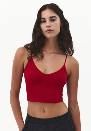 Mujer con cabello largo y oscuro, vestida con un top corto rojo y pantalones negros, sobre un fondo blanco, mirando al frente con una expresión neutral.