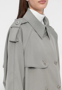 Trench coat grigio chiaro con colletto strutturato, design doppiopetto e spalline. Presenta una trama di tessuto e dettagli con bottoni.