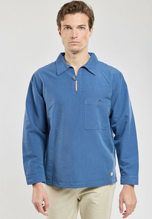 Homme portant un pull bleu à manches longues avec col boutonné et poche poitrine, debout devant un fond clair uni.
