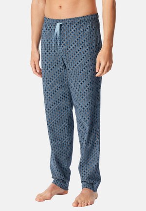 Homme adulte portant un pantalon de pyjama bleu à motifs avec un cordon de serrage bleu clair, debout pieds nus sur un fond blanc.