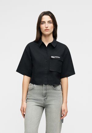 Žena v čiernej krátkej košeli s volnými rukávmi a predným vreckom, v kombinácii s vysokým pásom svetlosivých džínsov.