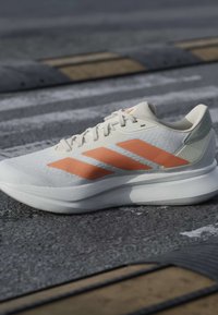 Chaussure de course blanche avec un marquage trois bandes orange sur un bitume gris près de ralentisseurs noirs et jaunes et de lignes de passage piéton blanches.