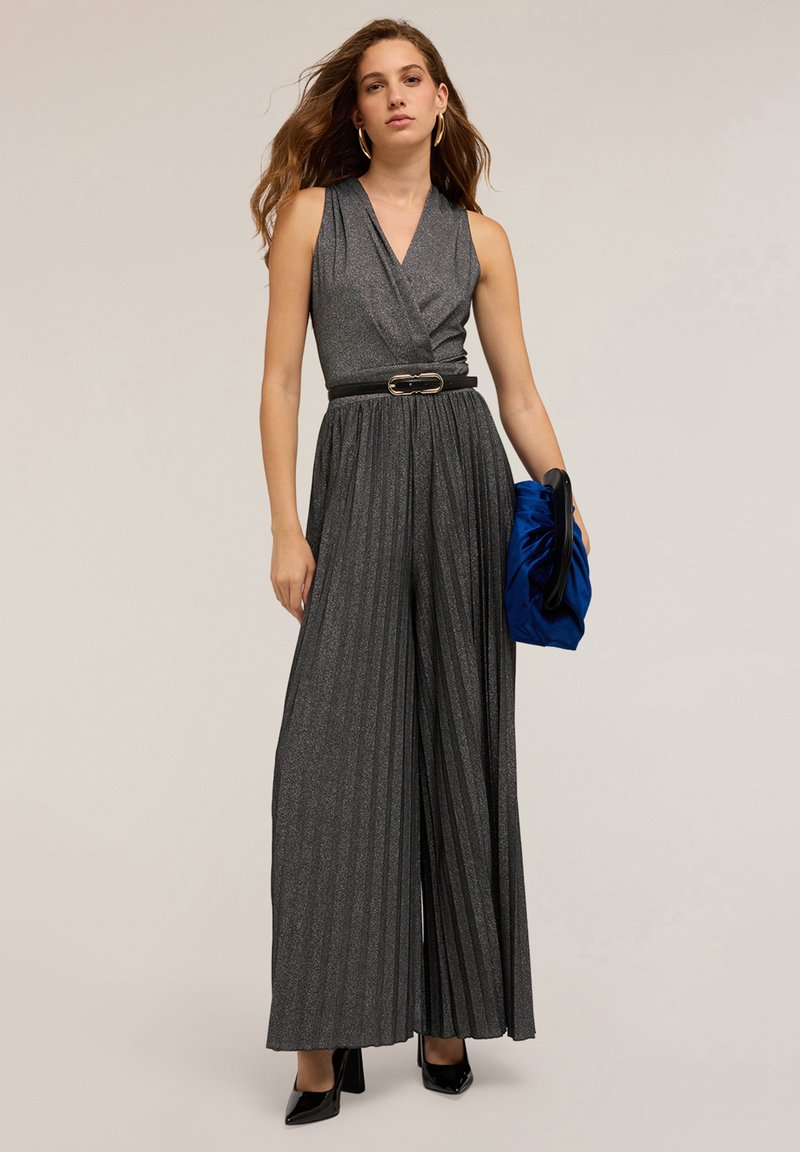 Jumpsuit grigio plissettato con scollatura profonda, vita cintrata e texture lucida, abbinato a una pochette blu e tacchi neri.
