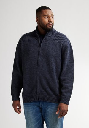 Jersey de punto navy con cremallera, con hombros acanalados texturizados, cuello alto y bolsillos laterales. Lleva puesto un top navy y unos pantalones vaqueros.