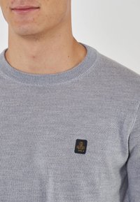 Maglione grigio a maglia con scollatura rotonda, caratterizzato da una piccola toppa nera con un emblema sul petto. Texture morbida e finitura del tessuto uniforme.