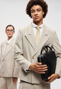 Fato bege sob medida com um blazer de dupla face, emparelhado com uma pasta de couro preto. O modelo tem cabelo encaracolado natural e uma gravata bege.