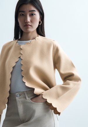 Νεαρή γυναίκα που φοράει μπεζ πουλόβερ με κυματιστό τελείωμα πάνω από λευκό μπλουζάκι και μπεζ παντελόνι, με το ένα χέρι στην τσέπη, ουδέτερο φόντο.