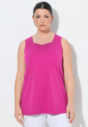 Kobieta z ciemnymi włosami zaczesanymi do tyłu, nosząca jasnoróżowy top bez rękawów z małymi ozdobami przy dekolcie oraz białe spodnie.