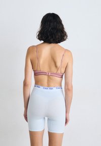 Una persona con capelli corti e ondulati è vista di spalle, indossando un reggiseno rosa e pantaloni corti bianchi Calvin Klein su uno sfondo chiaro.