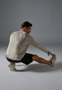 T-shirt d'entraînement beige clair à manches longues avec une texture en maille, ligne d'accent gris dans le dos, associé à un short noir et des chaussures d'athlétisme beiges.