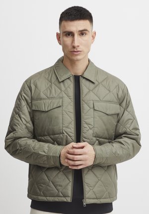 Quilted olive green jacket with a straight cut, featuring a collar, two chest pockets, and a front zipper. Smooth texture and lightweight material.