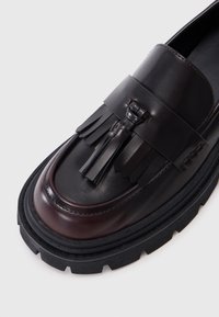 Loafers din piele cu o culoare maro închis, detalii cu ciucuri și o talpă groasă din cauciuc negru. Textură netedă cu accente contrastante.