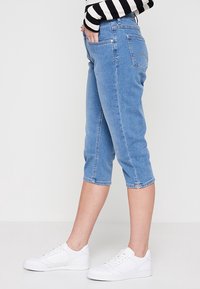 Jeans capri de mezclilla en azul claro con un diseño ajustado, bolsillos laterales y detalles de costura sutiles, combinados con zapatillas blancas.