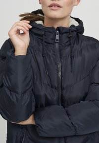 Marineblaue Pufferjacke mit Reißverschluss, Kordelzugkapuze und weichem Futter. Das Design beinhaltet diagonale Steppmuster und elastische Bündchen.