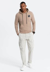 Felpa beige con cappuccio e tasca frontale, decorata con un logo, abbinata a pantaloni cargo chiari con diverse tasche. Sneakers bianche.