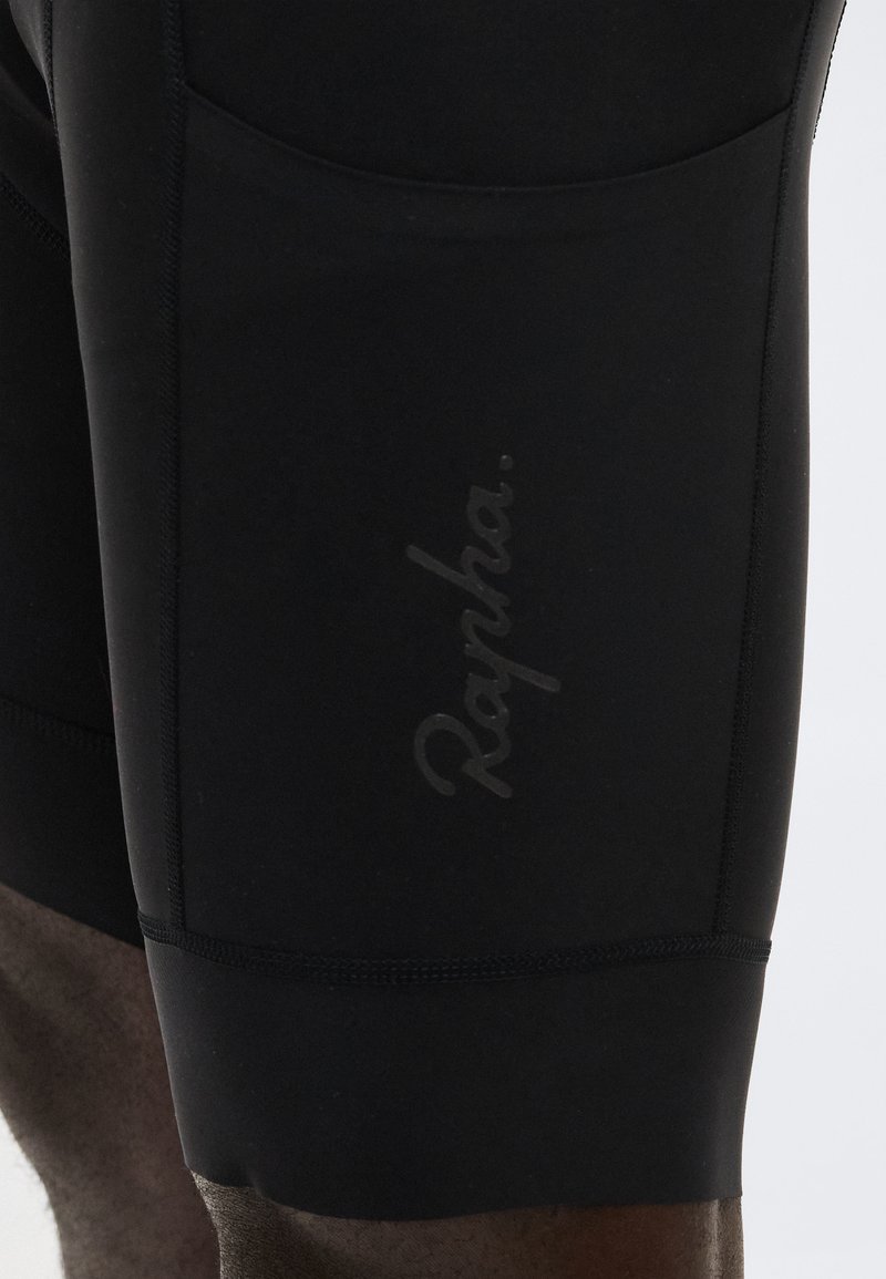 Close-up of black cycling shorts with subtle "Rapha" logo printed on the thigh, showing fabric texture and stitching details.
