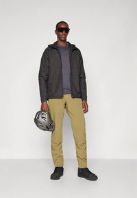 Sort letvægtsjakke, grå skjorte og beige bukser. Iført sorte sneakers og solbriller, holder en grå cykelhjelm og står foran en hvid baggrund.