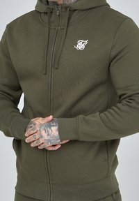 Sudadera con cremallera verde oliva hecha de tela suave, con capucha ajustable por cordón y logo bordado blanco en el lado izquierdo del pecho.