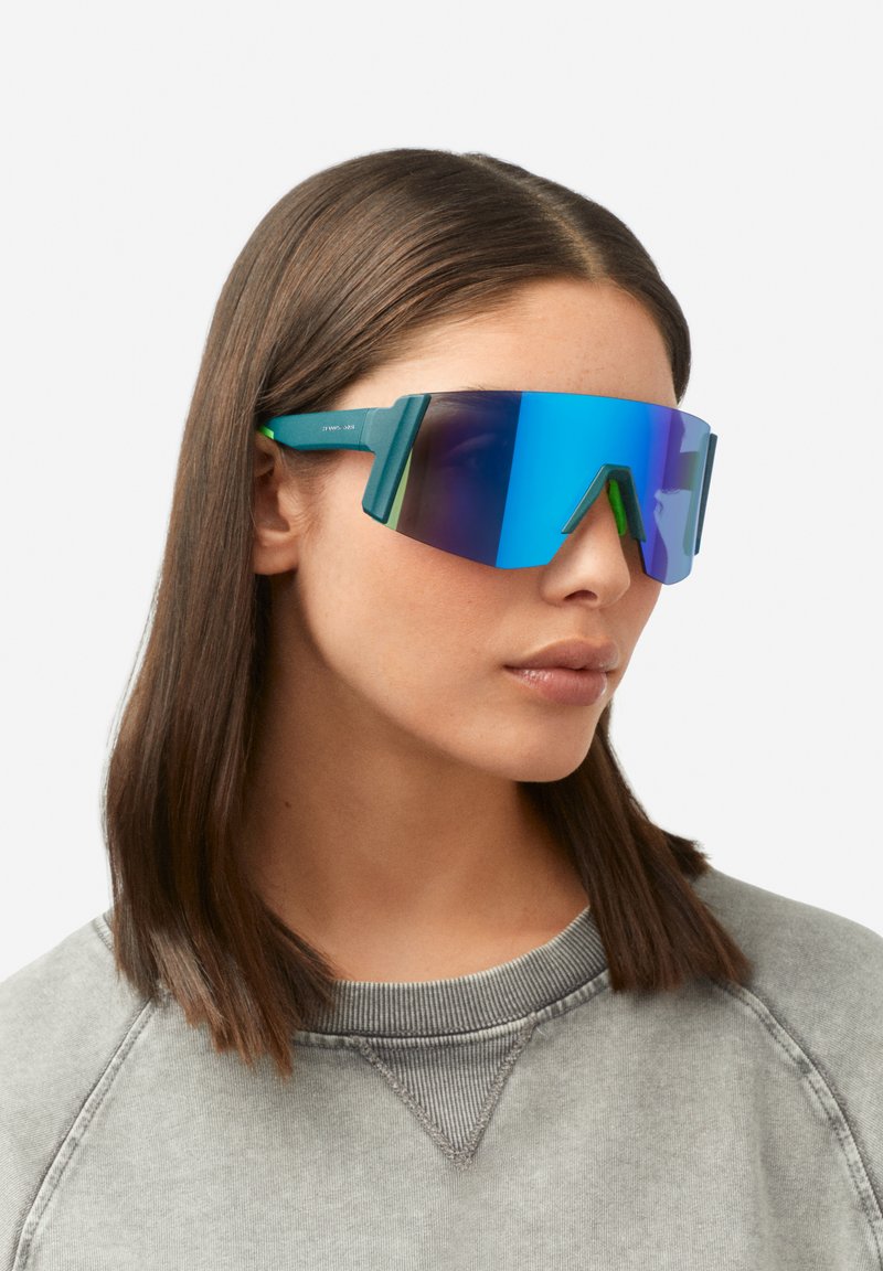 Zonnebril met een gedurfd, oversized ontwerp; blauwe spiegelglazen; matgroen montuur met geometrische accenten; lichtgewicht materialen.