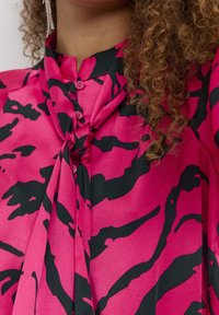 Różowa koszula z czarnym abstrakcyjnym wzorem, z detalem w postaci krawata i wieloma guzikami. Tkanina wydaje się gładka i lekka.