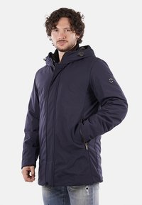 Giacca isolante blu navy con cappuccio, tasche con cerniera e una texture liscia. Presenta una patch con logo sul manica e chiusure a scatto.