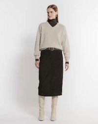 Kvinna står iförd en beige V-ringad tröja över en svart polotröja, en svart midikjol och beige knähöga stövlar framför en plain bakgrund.