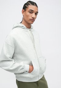 Svetlo siva zip-up kapuca s kapuljačom na vezanje, prednjim žepovima i rebrastim manžetama, izrađena od mekog, teksturiranog materijala.