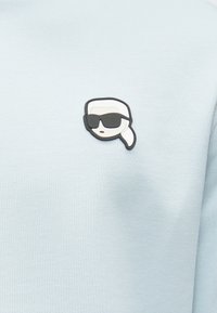 Felpa azzurro chiaro con una toppa in gomma raffigurante un volto con coda di cavallo e occhiali da sole, posizionata sul lato sinistro del petto.