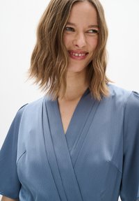 Blaue Wickelkleid aus strukturiertem Stoff, mit kurzen Ärmeln und tiefem V-Ausschnitt. Besitzt klare Linien und einen lockeren Schnitt für zusätzlichen Komfort.
