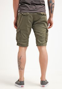 Pantalones cortos de cargo verde oliva con bolsillos laterales, hechos de una tela ligera. Presentan puños terminados y un ajuste relajado. Llevados con zapatillas grises.