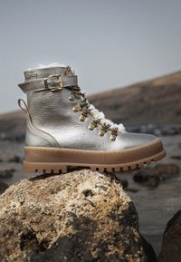 Botín plateado metálico con forro de pelo, diseño de cordones, herrajes dorados, punta redonda y suela marrón rugosa con un dibujo pronunciado.