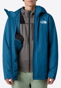 Uomo che indossa una giacca blu The North Face con cappuccio, una felpa grigia con zip e pantaloni verde chiaro, mentre tiene un dispositivo nero nella tasca interna della giacca.