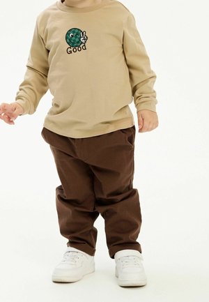 Tout-petit portant une chemise à manches longues beige avec un motif de visage souriant vert et signe de paix, un pantalon marron et des baskets blanches, debout sur un sol blanc.