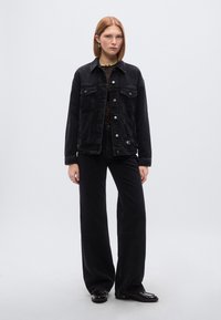 Μαύρο denim μπουφάν με τσέπες στο μπροστινό μέρος, ασημένια κουμπιά και άνετη εφαρμογή, συνδυασμένο με μαύρο παντελόνι φαρδιάς γραμμής και μαύρα παπούτσια.