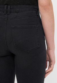 Melna džinsu bikses ar augstu jostasvietu, ar aizmugures kabatu un smalkiem šuvuma detalēm. Audumam ir gluda tekstūra.