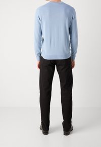 Maglione a maglia azzurro chiaro con polsini e orlo a costine, indossato con pantaloni neri e scarpe scure, visto da dietro su uno sfondo neutro.