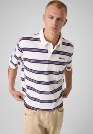 Jeune homme portant un polo blanc à rayures horizontales bleu marine et rouges et un pantalon beige, debout devant un fond clair uni.