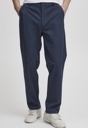 Homme portant un pantalon bleu marine et des baskets blanches, debout les mains dans les poches devant un fond uni.
