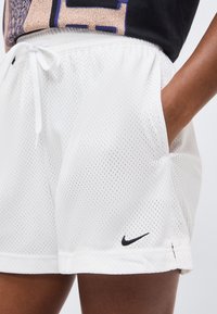 Άσπρα αθλητικά σορτς από δίχτυ με ελαστική μέση και κορδόνι, χέρι στην τσέπη, και μαύρο λογότυπο Nike Swoosh στο αριστερό πόδι.