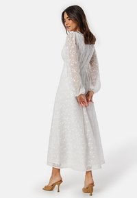 Vit maxiklänning med blommönster, långa genomskinliga ärmar, cinchad midja och svängig siluett; bärs med ljusbruna klacksandaler.