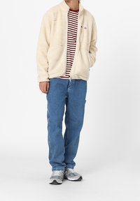 Giacca in pile beige con colletto alto e due tasche frontali, indossata sopra una maglietta a righe rosse e bianche, con jeans blu e sneakers grigie.