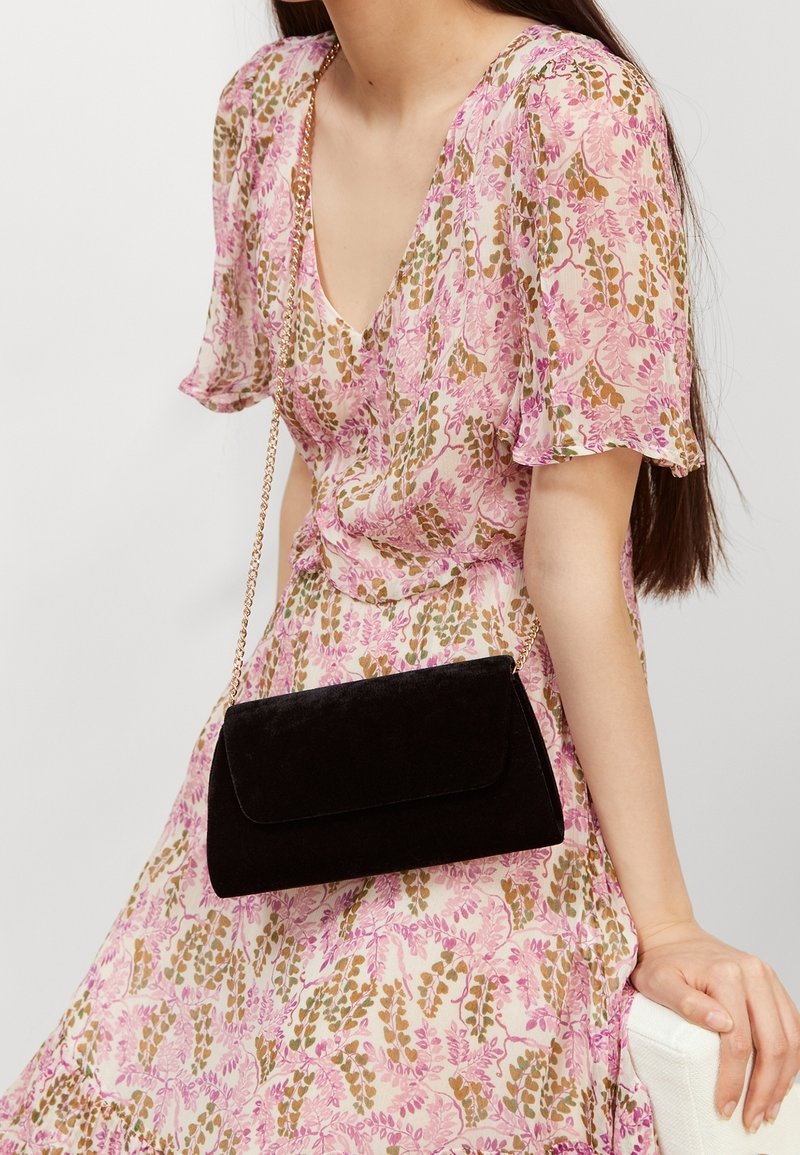 Sort velour clutch med magnetisk flaplukning, prydet med en guldkædetræ. Sat mod en blomstret kjole i pink og grønne nuancer.
