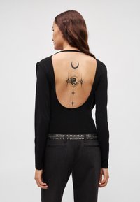 Czarna bluzka z długim rękawem z głębokim wycięciem na plecach, zestawiona z ciemnymi, dopasowanymi spodniami z ćwiekowanym paskiem. Na plecach widoczny tatuaż z czarnego tuszu.