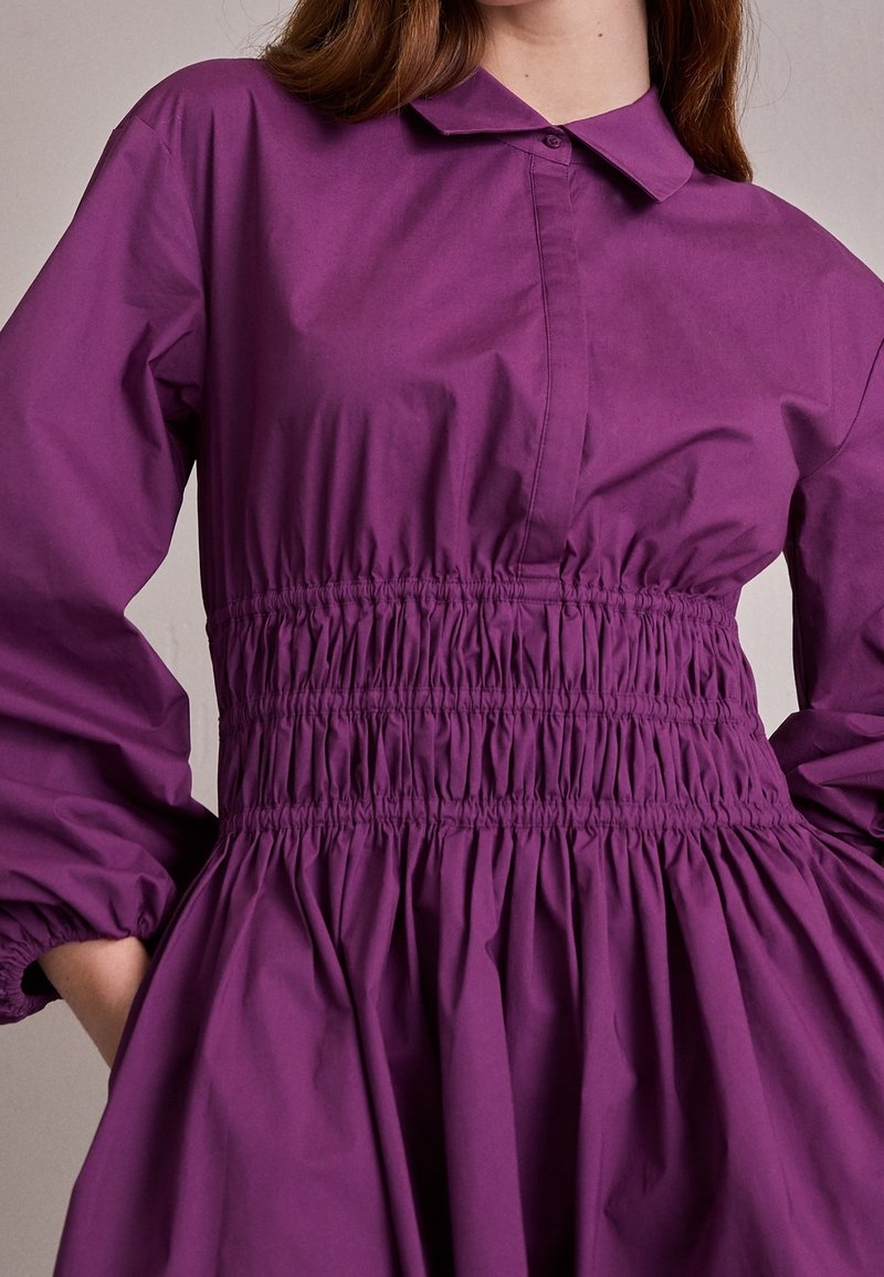 Robe violette avec une taille froncée, dotée d'un col et de manches longues bouffantes. Confectionnée dans un tissu lisse et léger.