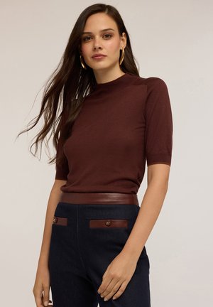 Donna con lunghi capelli castani, che indossa orecchini a cerchio dorati, una maglietta corta color marrone scuro e pantaloni scuri con tasche rifinite in pelle e cintura.