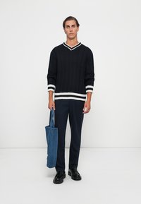Jeune homme en pull noir à rayures blanches, pantalon bleu marine, chaussures noires, tenant un sac fourre-tout bleu, se tenant contre un mur blanc.