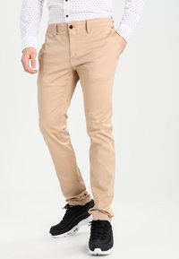 Beige chinos made from cotton with a slim fit, featuring front pockets and a button closure, paired with black sneakers.