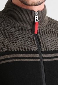 Zbliżenie na mężczyznę w brązowym i czarnym dzianinowym swetrze zapinanym na zamek z czerwonym uchwytem w kształcie litery B.