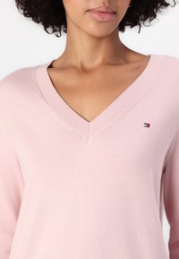 Roza pulover z V-izrezom iz mehkega pletenega blaga z rebrasto obrobo. Ima majhen vezeni logotip v rdeči, beli in mornarski modri barvi na prsih.