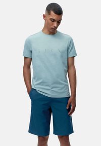 Svijetloplava majica kratkih rukava s suptilnim grafičkim dizajnom na prednjem dijelu, uparen s tamnoplavim šortsevima s bočnim džepovima.